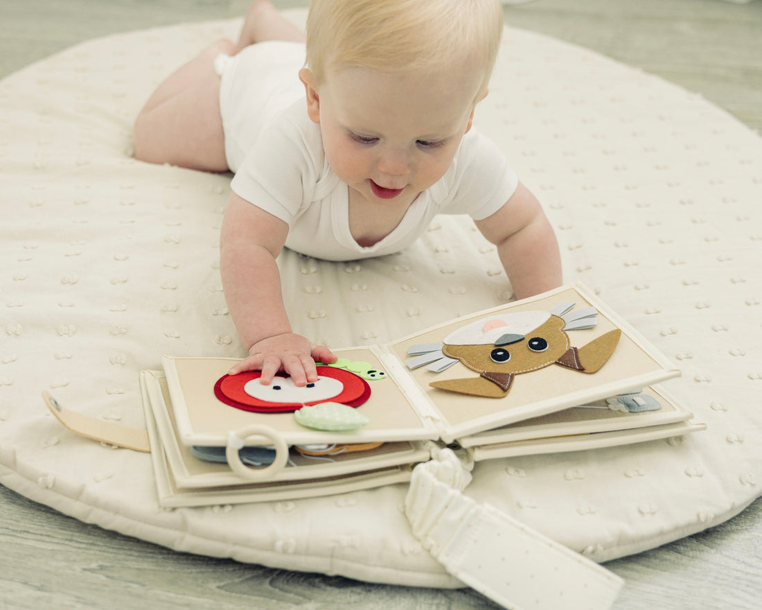 Baby lying on a soft beige mat playing with a Montessori felt busy book featuring interactive shapes and textures, promoting fine motor skills and sensory learning.