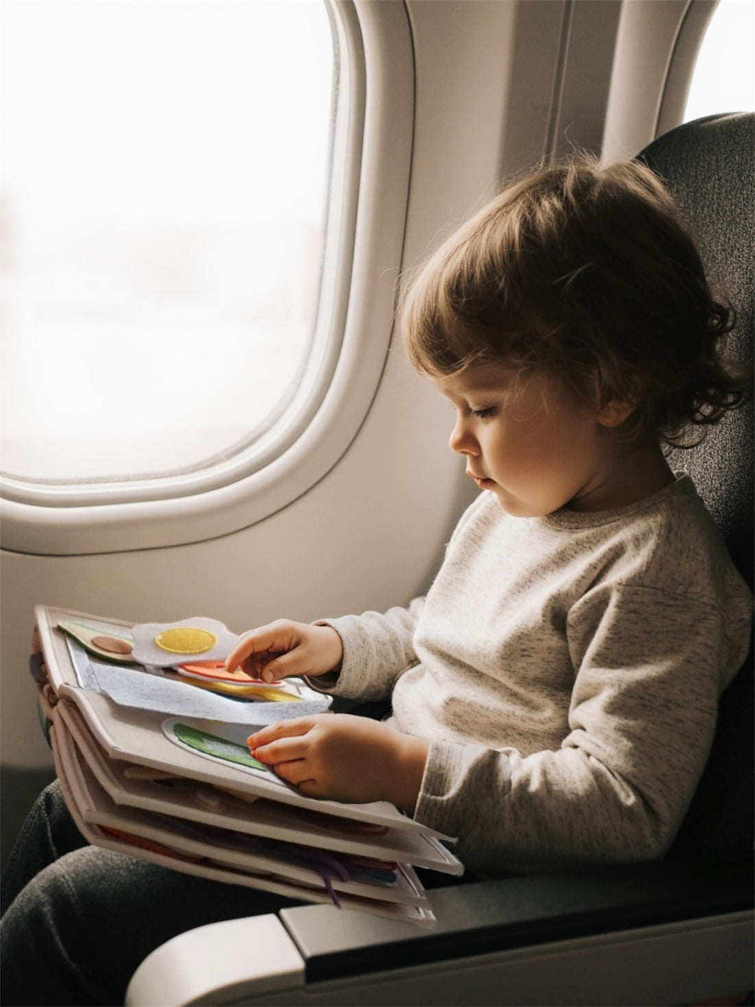 A toddler sitting by an airplane window, quietly playing with a soft felt activity book during a flight.